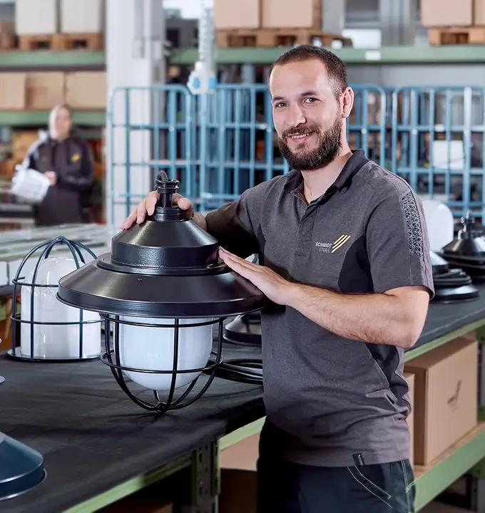 ein Schmidt-Strahl Mitarbeiter in der Produktion hält eine Straßenleuchte in der Hand, umgeben von weiteren Leuchten und Arbeitsmaterialien auf einer Werkbank