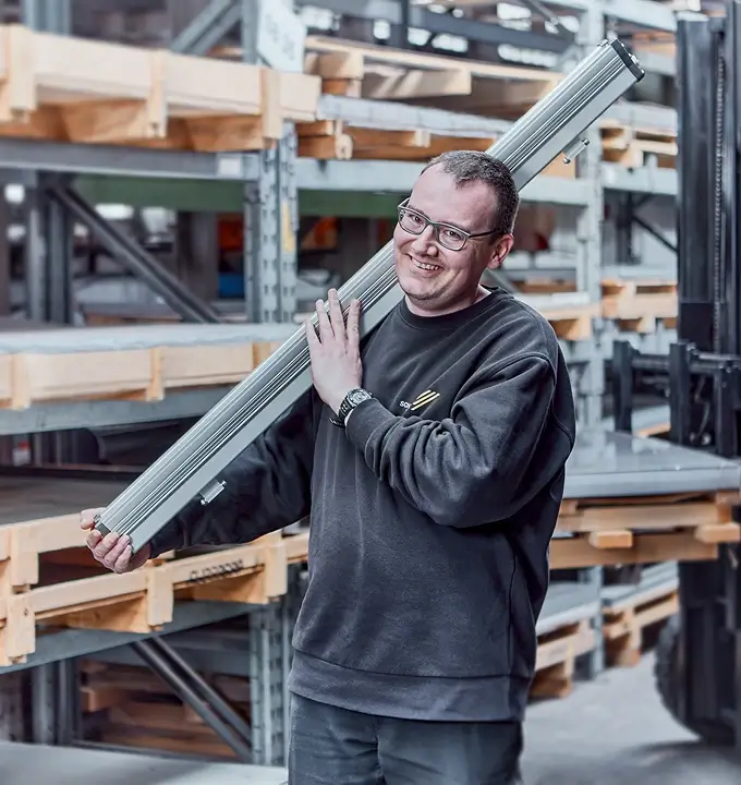 Mitarbeiter von Schmidt-Strahl trägt eine lange LED-Leuchte vor Lagerregalen in der Fertigung.